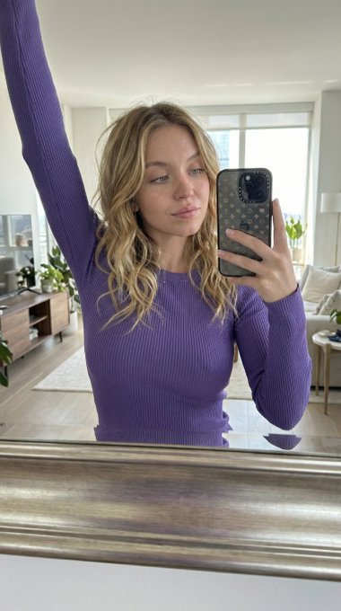 A woman taking a mirror selfie with a soft confident expression, relaxed posture, holding a smartphone in a neutral-toned room.
