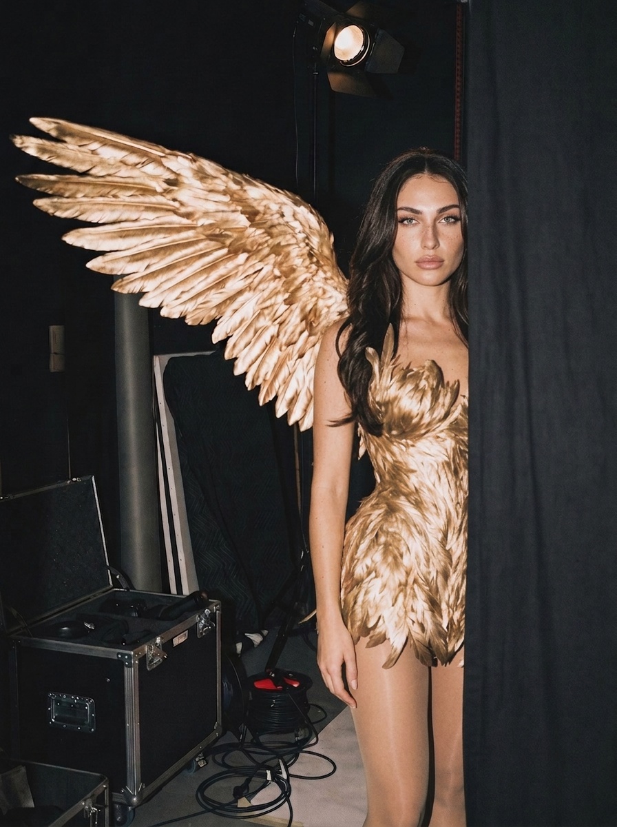 A person in a golden feathered bodice with large wings, standing in a studio with a black curtain, illuminated by overhead light.