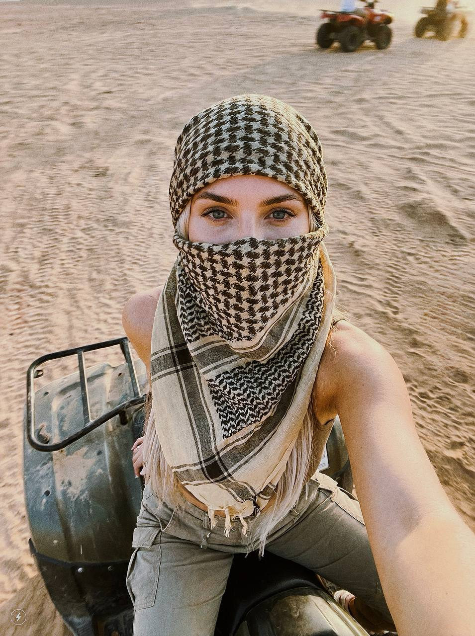 A woman in a sandy desert, seated on a quad bike, with a scarf covering her lower face, eyes visible, in warm light.