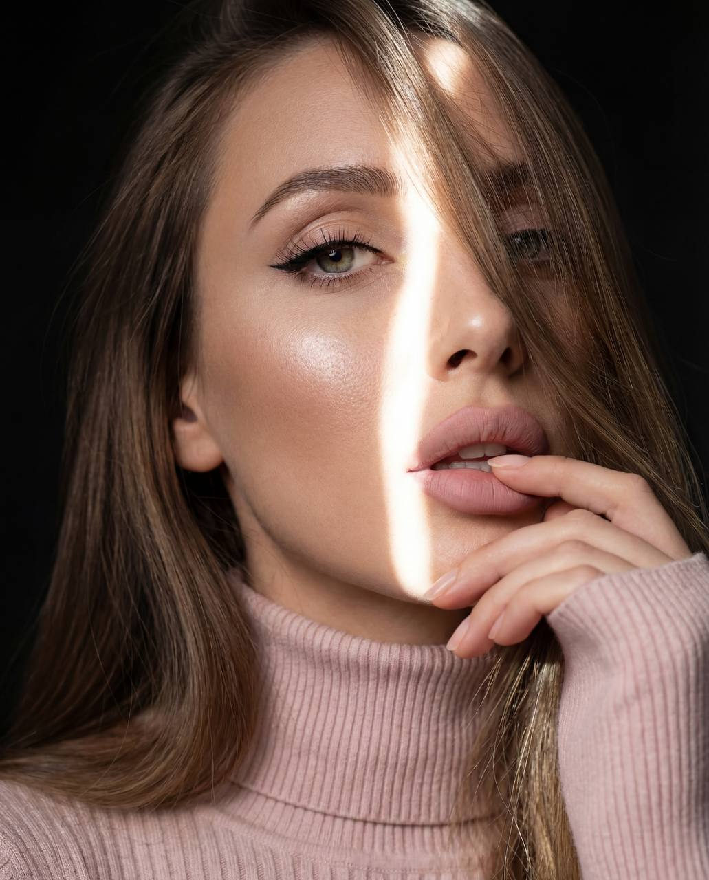 Close-up portrait of a young woman with long hair cascading down her face, illuminated by a soft light strip, wearing a pale pink sweater.