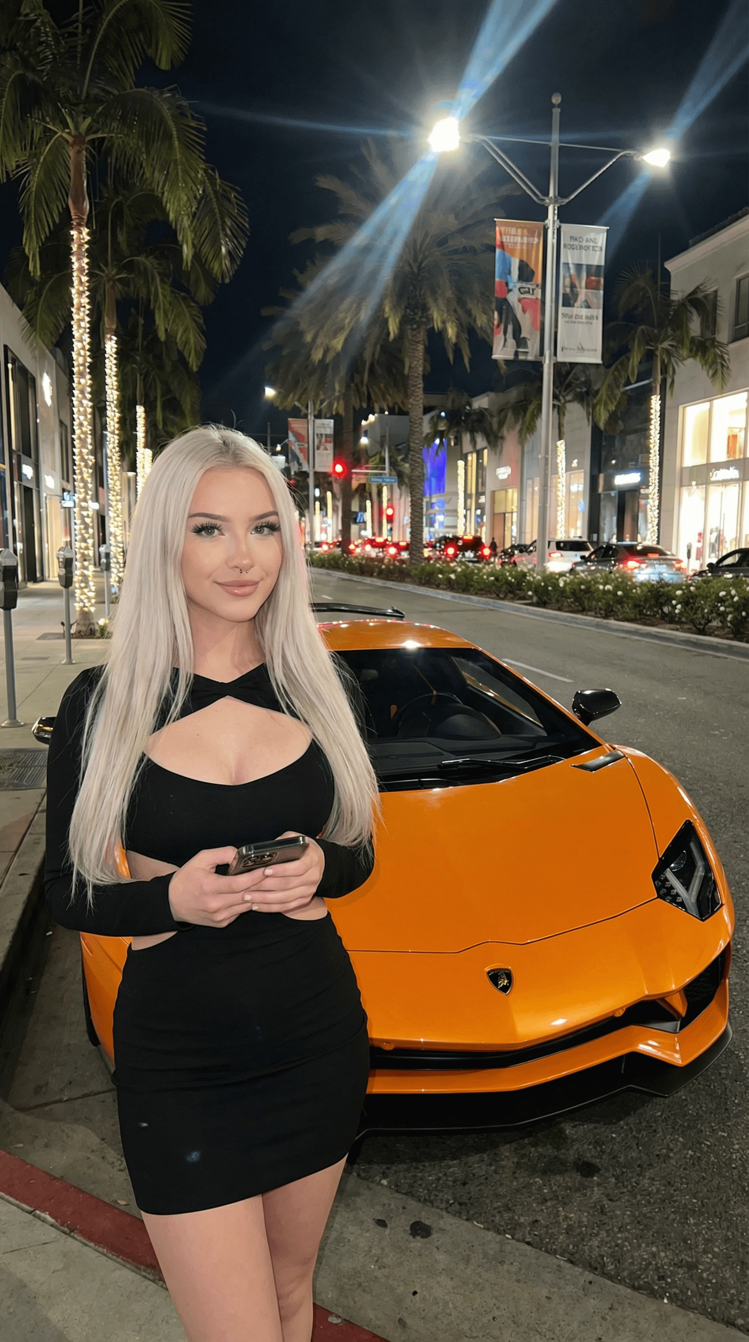 A model in a black dress stands beside an orange Lamborghini Aventador on a beautifully lit city street at night.