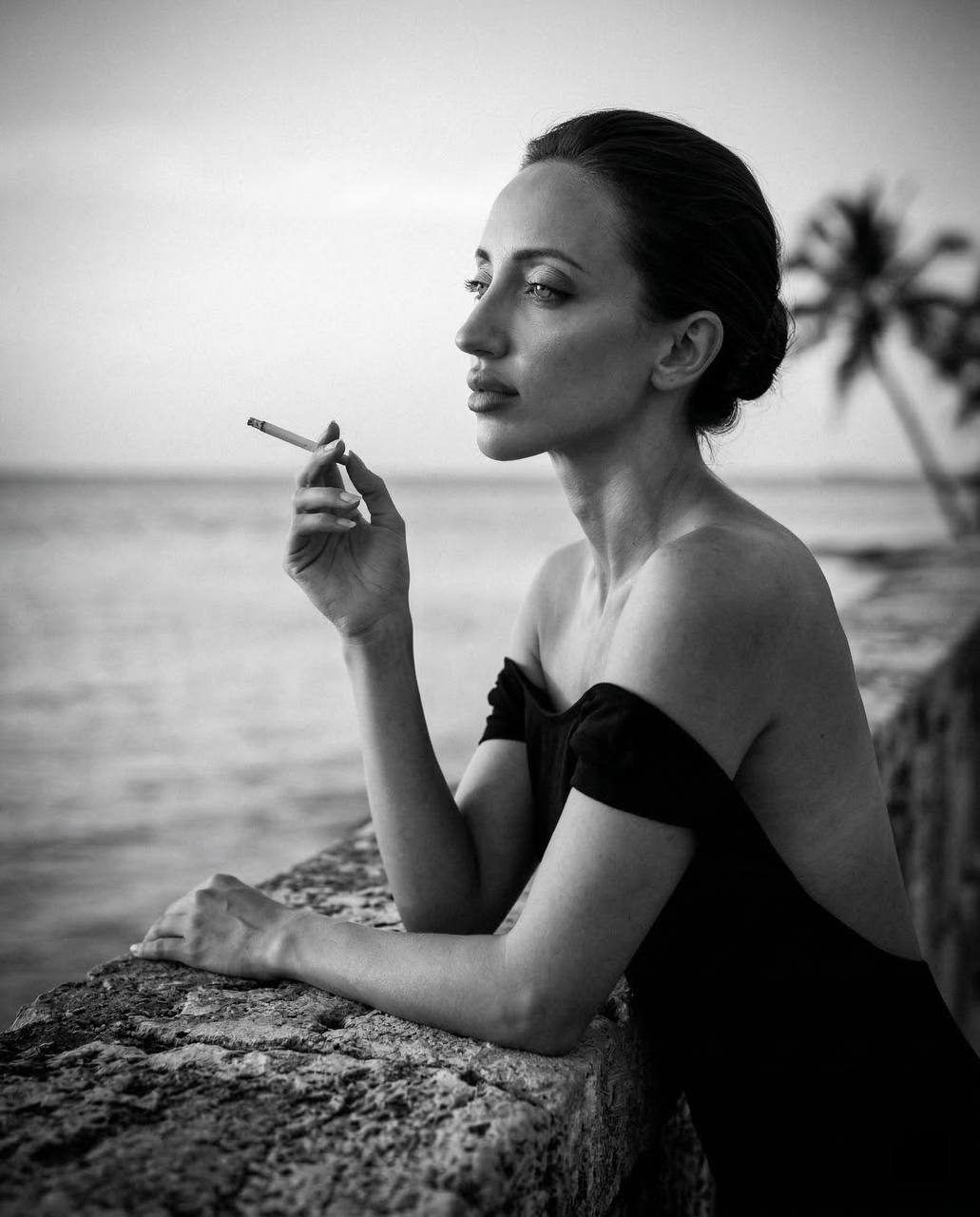An elegant girl in profile at the seashore, lean on a stone parapet with a cigarette. Monochrome with high contrast.
