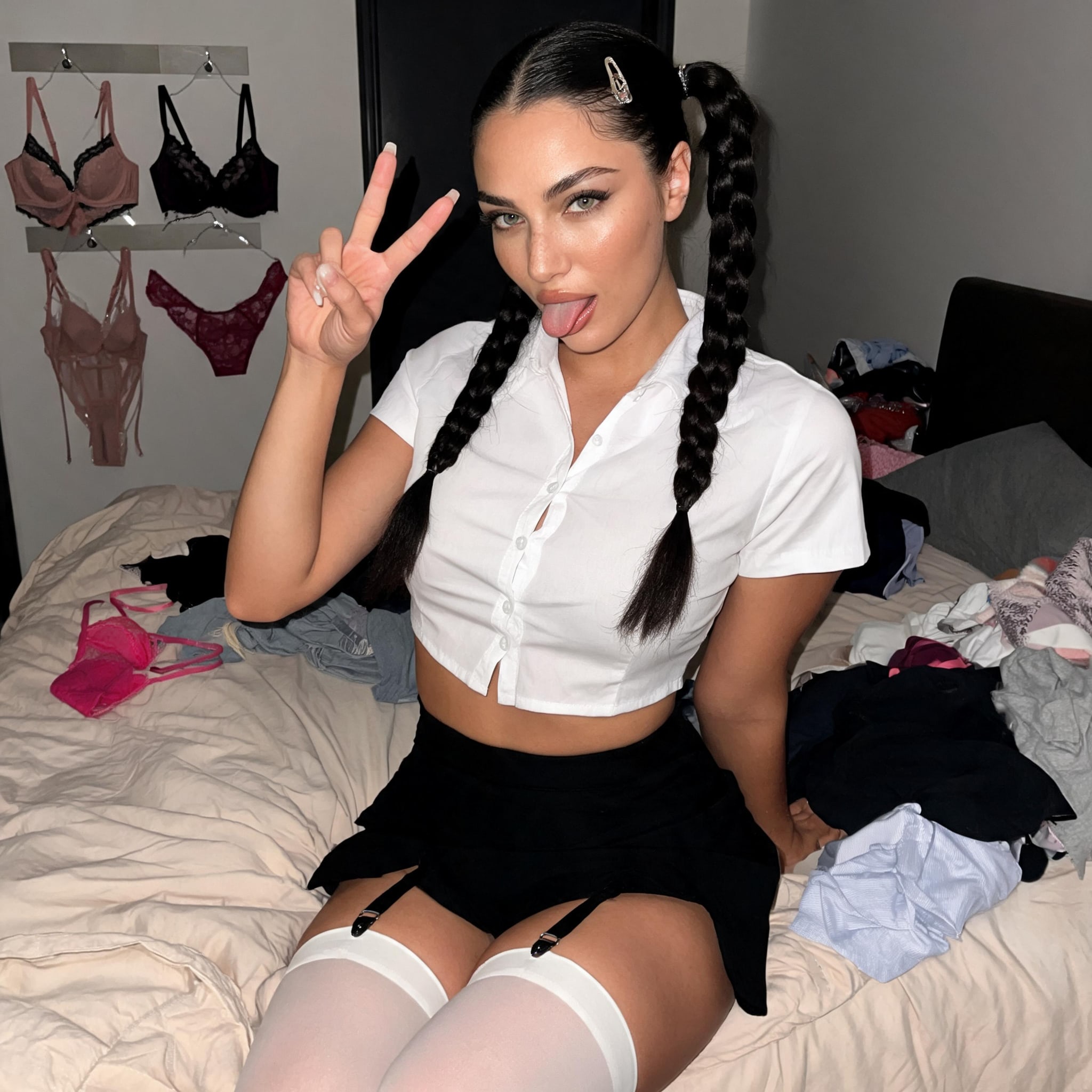 A woman wearing a cropped white blouse and black mini skirt, playfully posing with a peace sign on a messy bed.