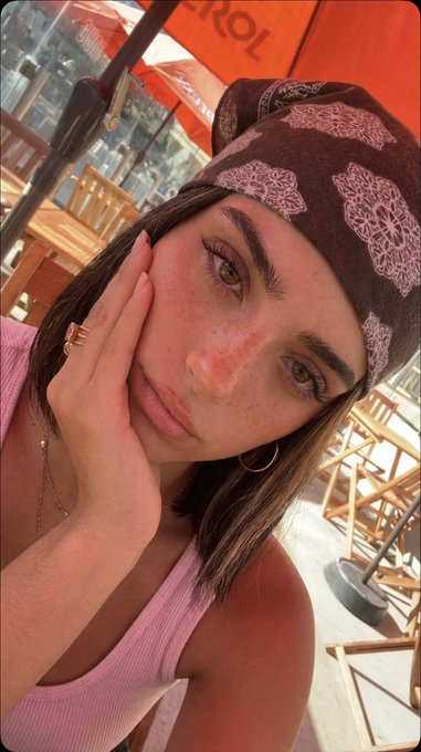 Close-up of a woman wearing a brown bandana and pink tank top in an outdoor setting, sunlight highlighting her features.