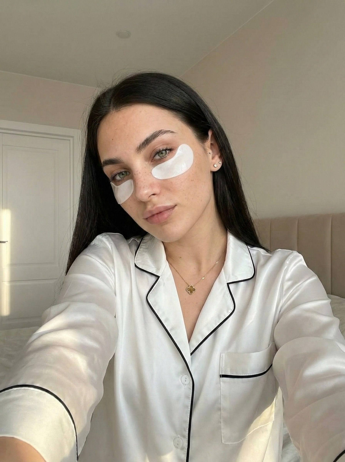A woman with long straight hair taking a selfie in a cozy bedroom, wearing satin pajamas and natural makeup, with hydrogel eye patches.