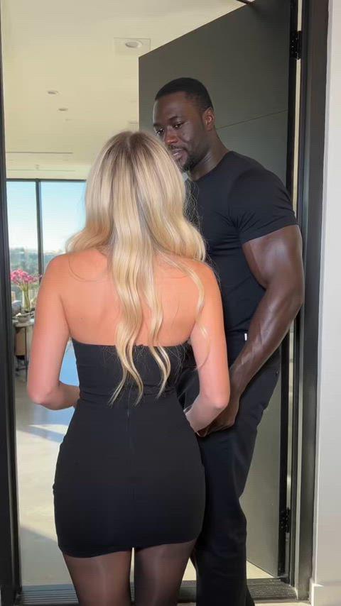 A woman in a tight black strapless mini dress stands in a modern apartment doorway with a muscular man.