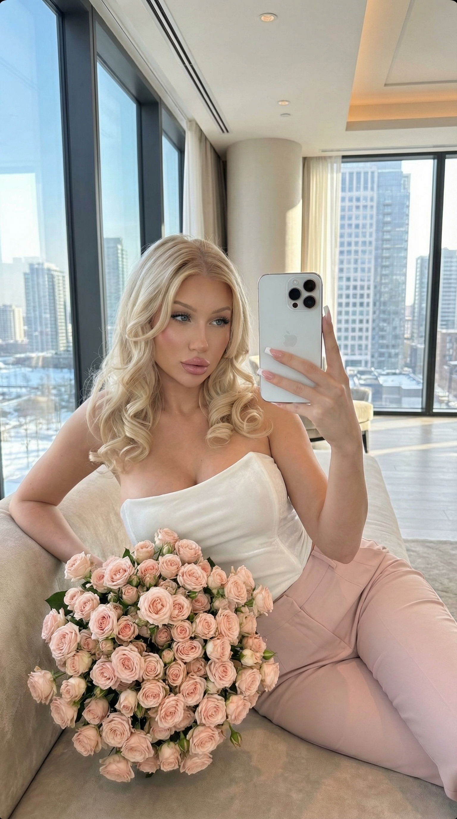 A woman in a white velvet top and pink trousers holds a bouquet of blush-pink roses while seated in a luxury hotel lobby, capturing a mirror selfie.