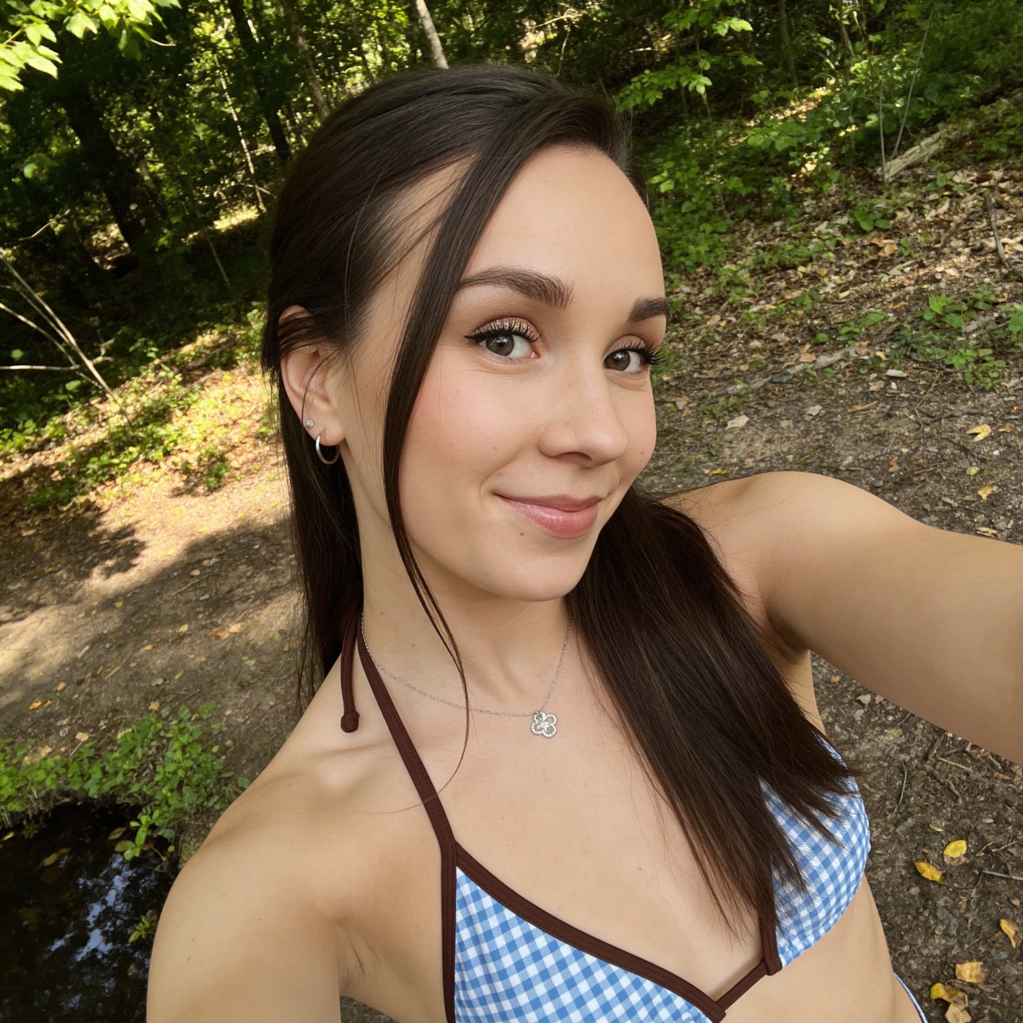 A woman with soft makeup and a blue-and-white gingham bikini top takes a selfie in a wooded area, sunlight filtering through the leaves.