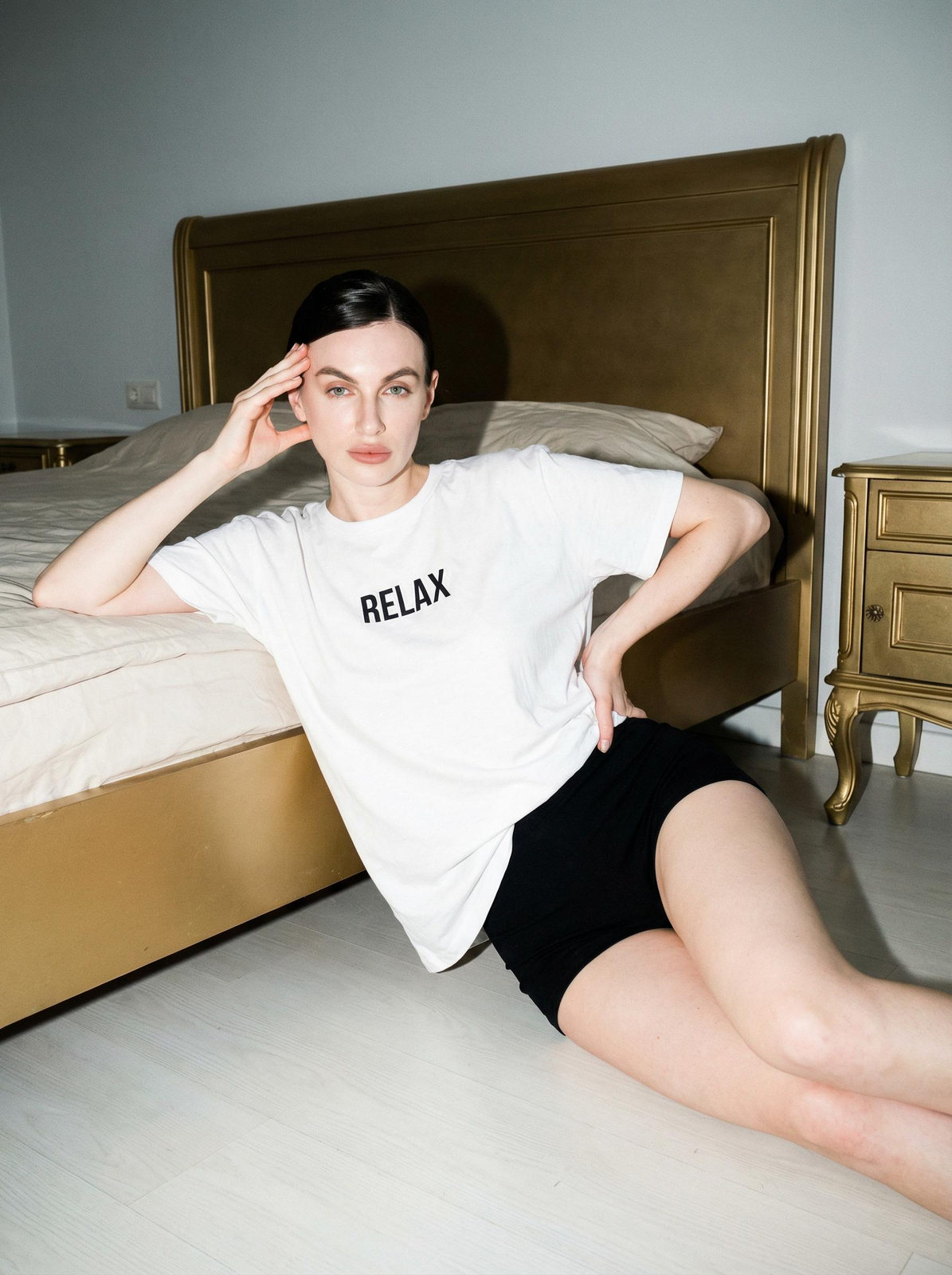 Woman reclining on the floor in a vintage bedroom wearing an oversized T-shirt and high-waisted shorts, looking directly into the camera.