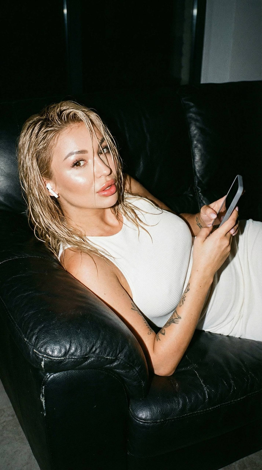 A girl reclining on a leather sofa in a realistic night flash portrait, wearing a ribbed dress and holding a gray phone.