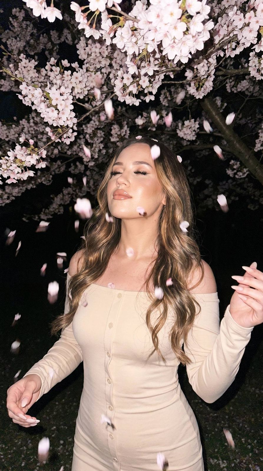 A woman under blooming cherry branches with long soft waves and a soft cream-chocolate dress, eyes closed in a calm dreamy pose.