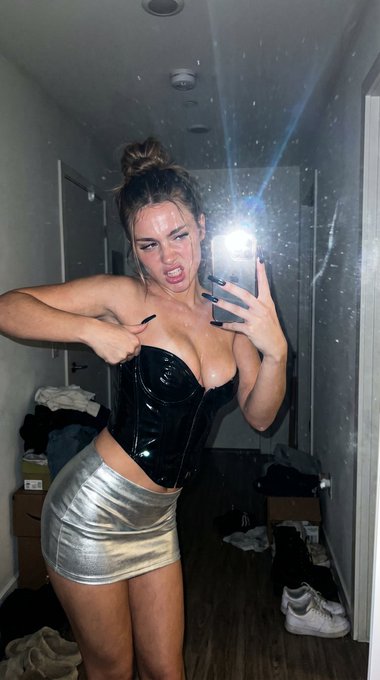 A woman takes a selfie in a mirror wearing a black patent corset and a silver metallic skirt, illuminated by a smartphone flash.