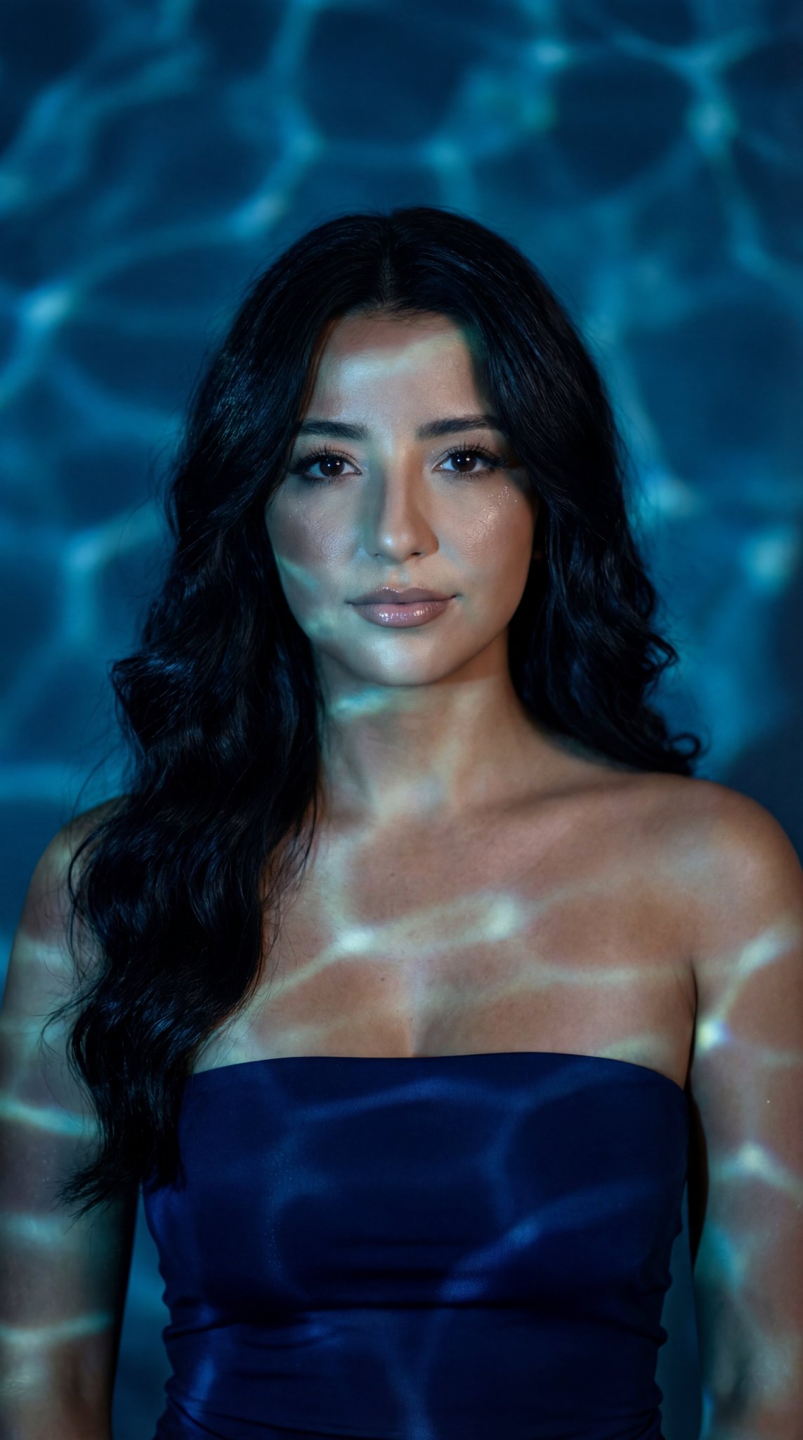 A female model in a deep navy strapless blouse with voluminous hair against a deep blue water ripple background.