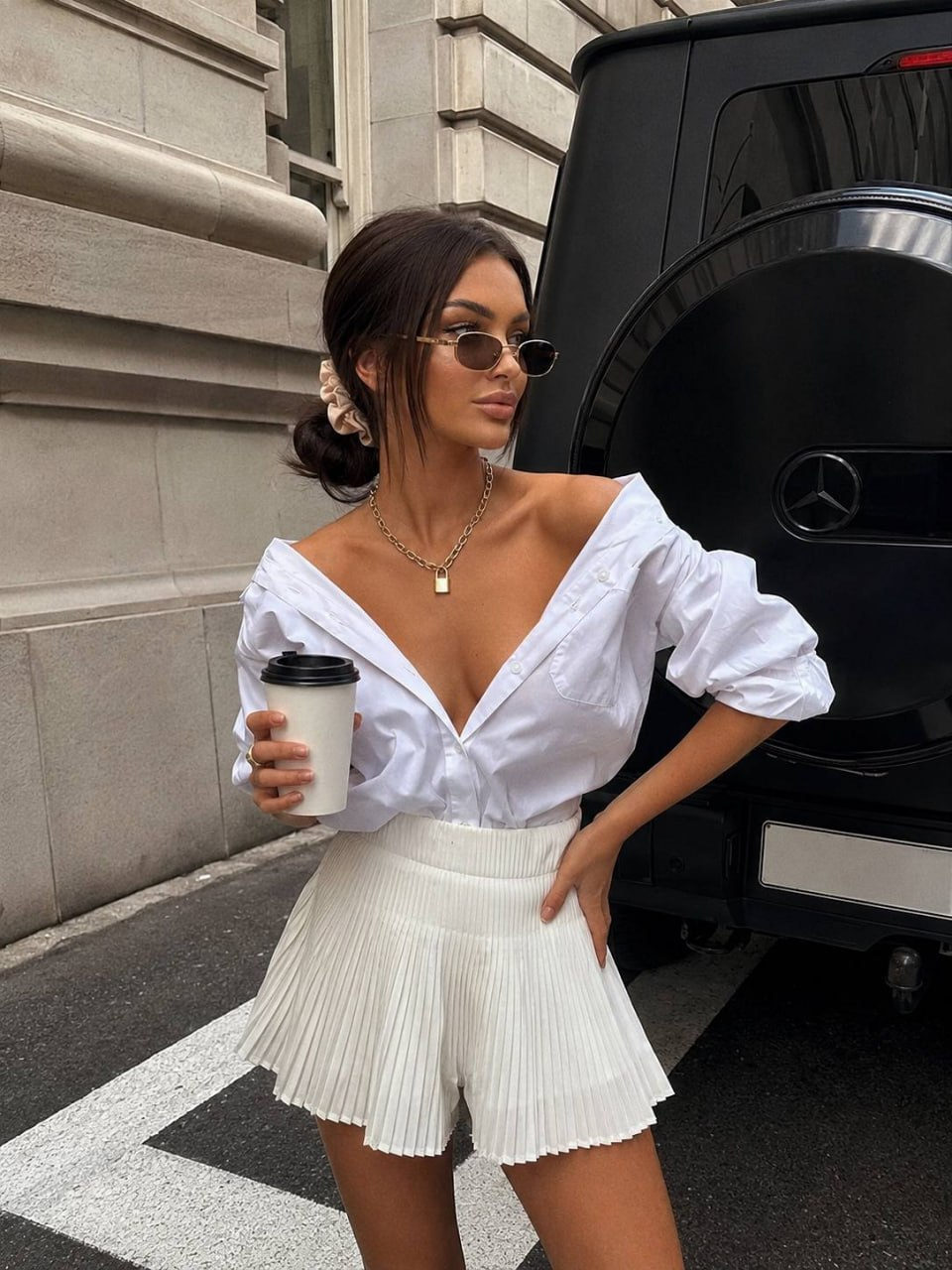 A woman in an off-shoulder white shirt and pleated skirt poses elegantly beside a black Mercedes-Benz G-Class SUV on a city street.