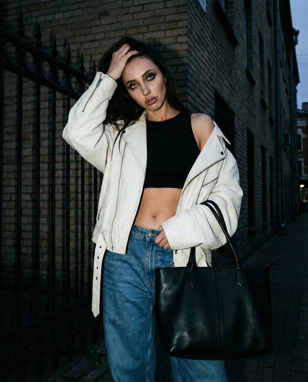 Close-up of a woman in an oversized vintage leather jacket, adjusting her hair against a dark city street background, with muted twilight lighting.
