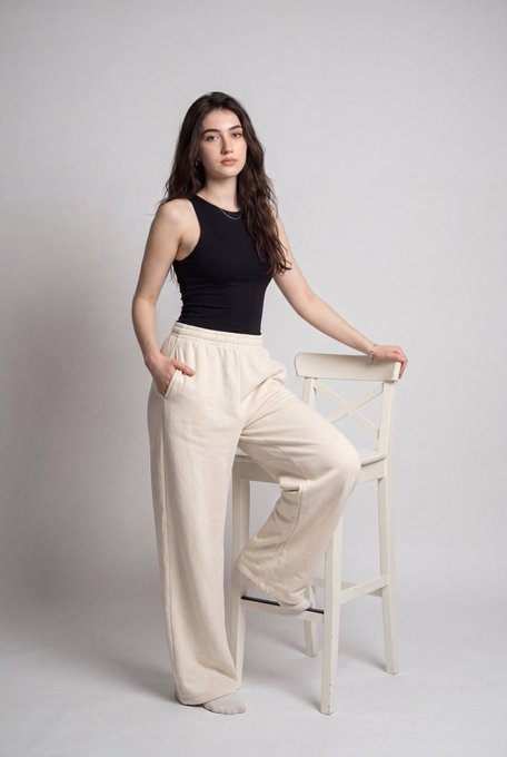 Full-body portrait of a confident woman in a black sleeveless tank top and light beige wide-leg pants, posed against a seamless light gray backdrop.