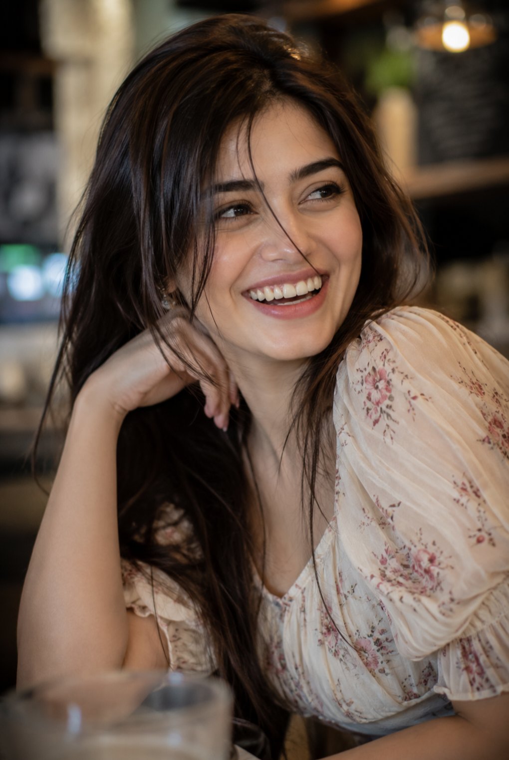 Close-up portrait of a woman mid-laugh, wearing a cream floral top with gathered puff sleeves in a warm indoor setting.