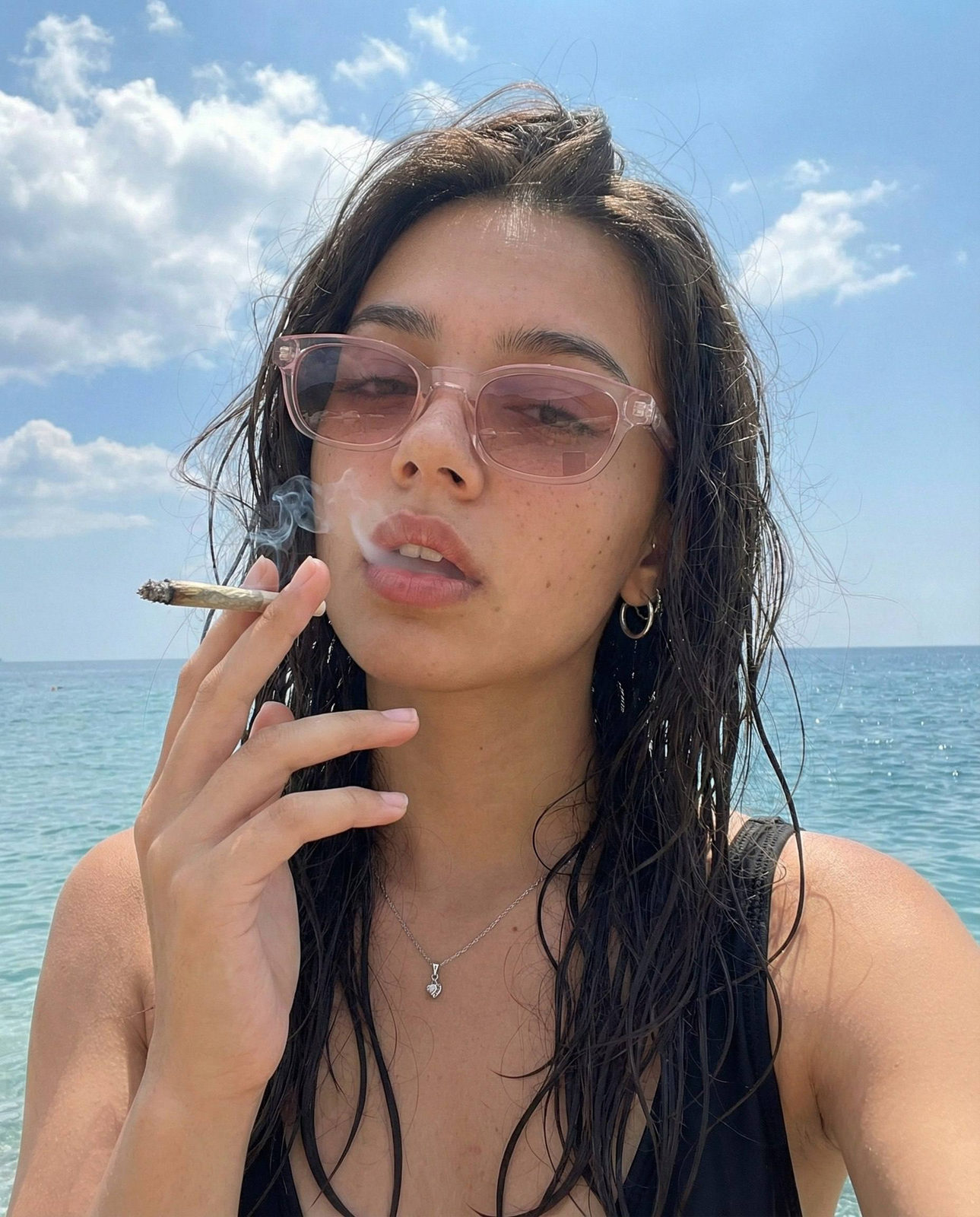 Young woman with wet hair taking a selfie at the beach, wearing black swimsuit, surrounded by blue sea and clouds.