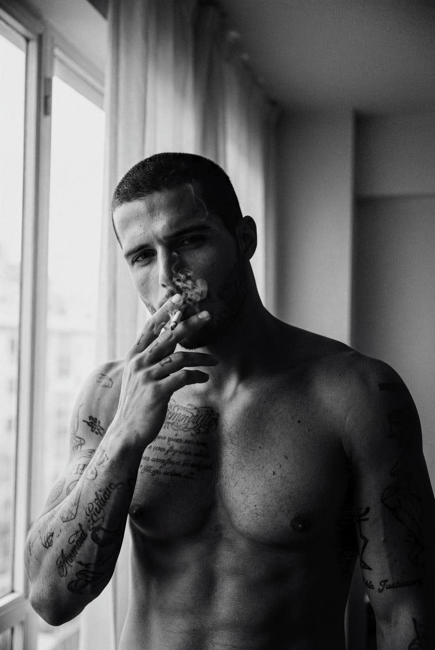 A shirtless man with tattoos stands indoors by a large window, exhaling softly with a calm expression.