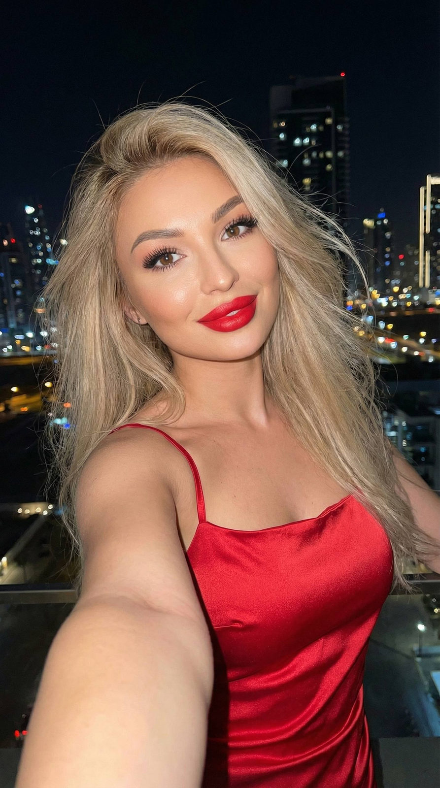 A girl in a bright red satin dress with thin straps stands in a city at night, her hair fluttering in the wind while she enjoys a playful moment.