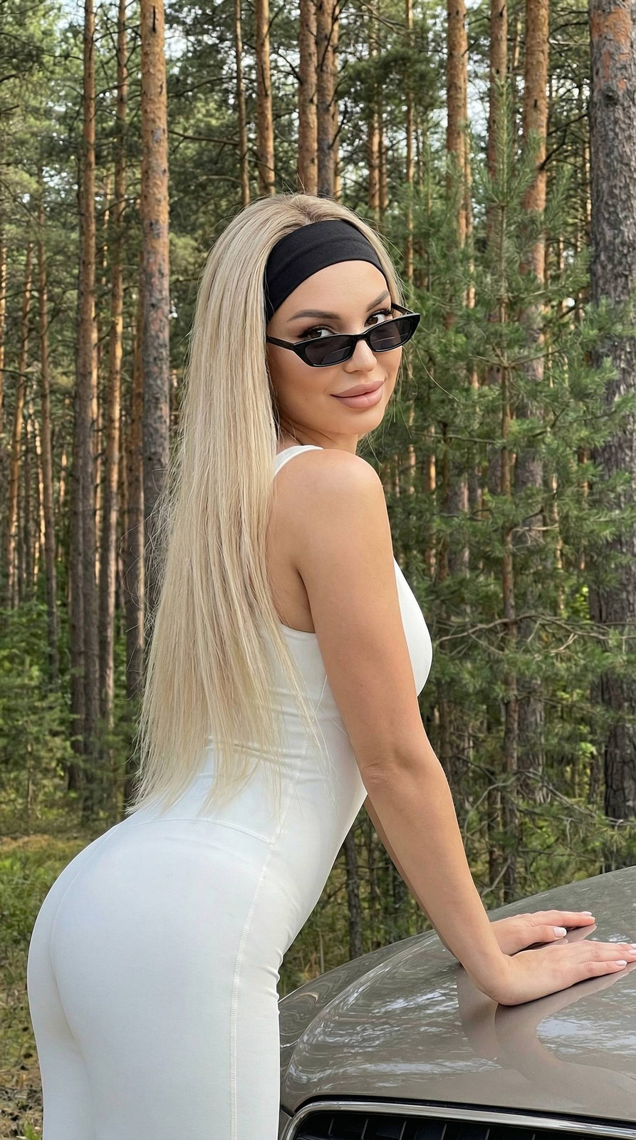 Girl in a white jumpsuit leaning on a car hood in a pine forest, back to the camera, with a slight smile and black sunglasses.