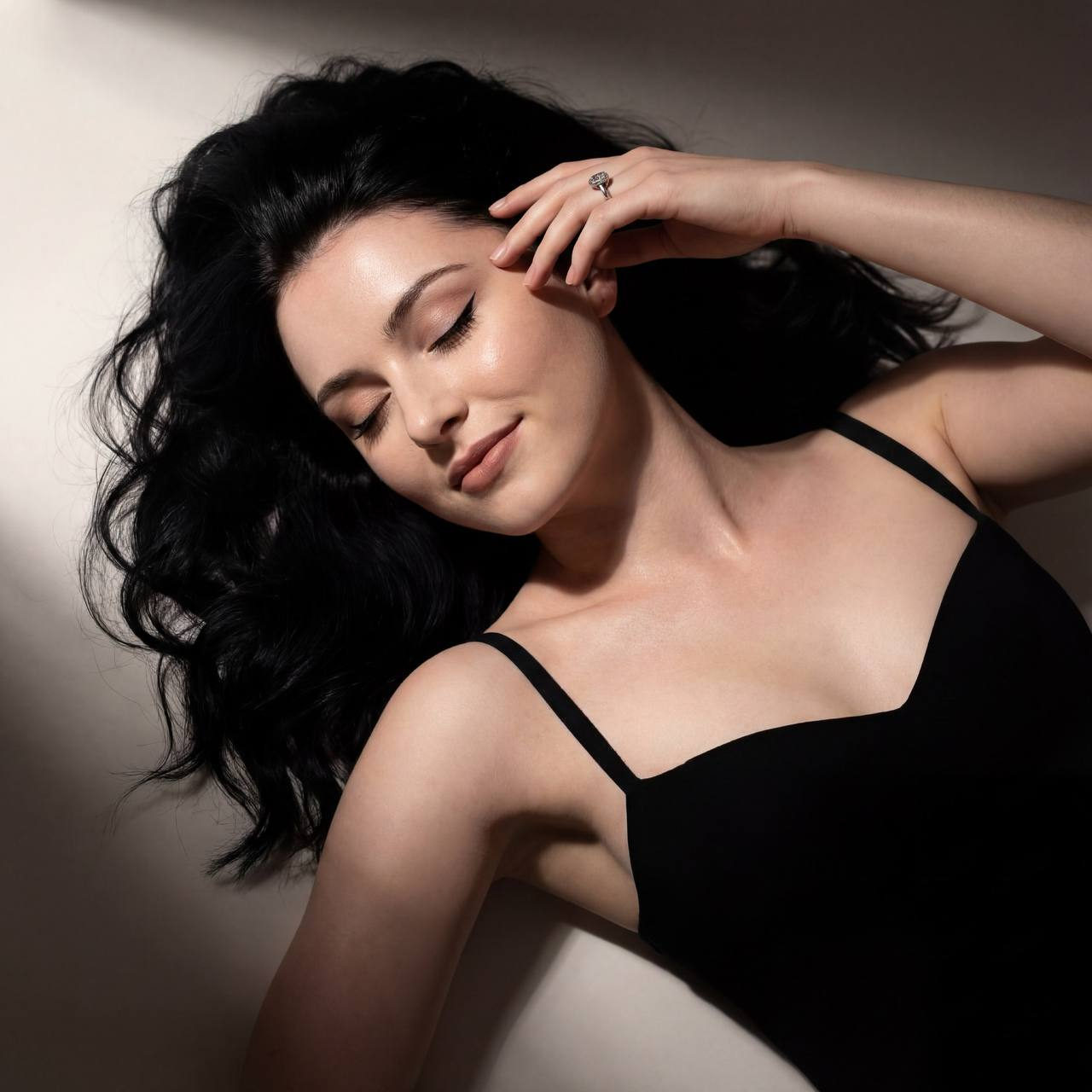 A woman in a form-fitting evening dress lying gracefully on a light surface, with long dark hair and a soft smile, captured in dramatic chiaroscuro lighting.