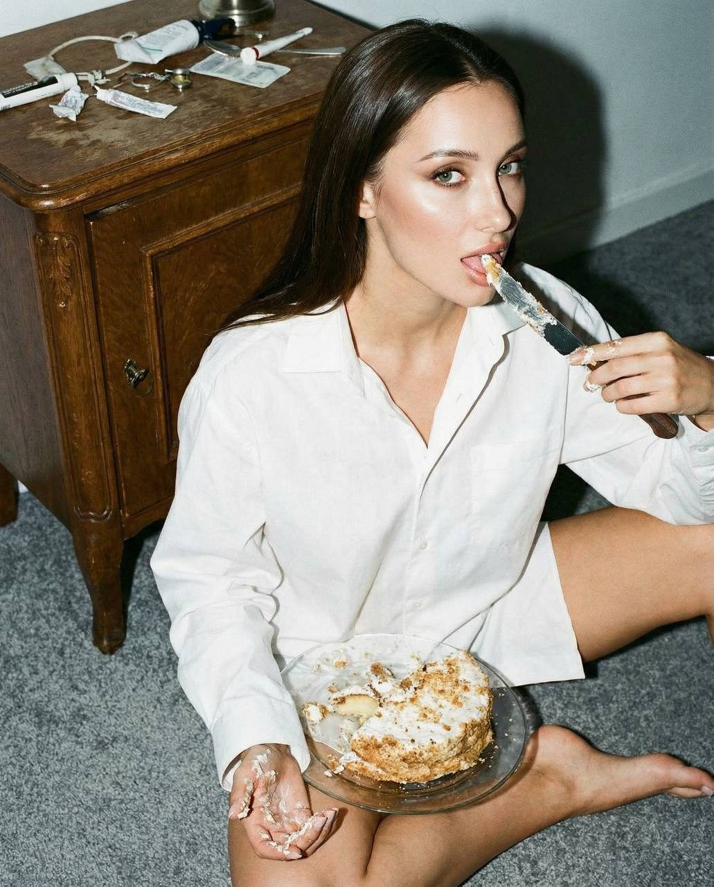 Cinematic close-up portrait of a woman sitting on the floor, wearing a men's shirt, with a bitten cake and knife.