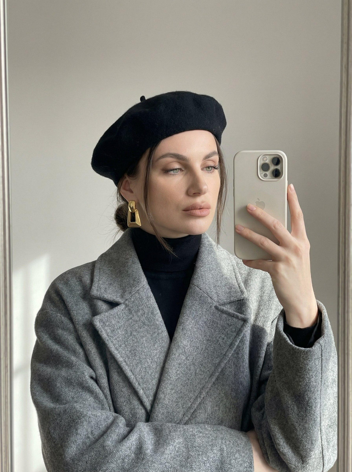 A woman in a gray coat standing in front of a mirror taking a selfie, with soft lighting highlighting her features.