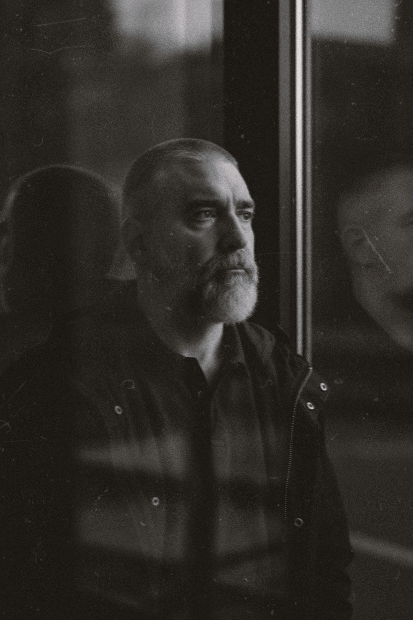 A middle-aged man with a gray beard stands by a window, wearing a dark polo and a black military jacket, captured in a soft, moody black and white.