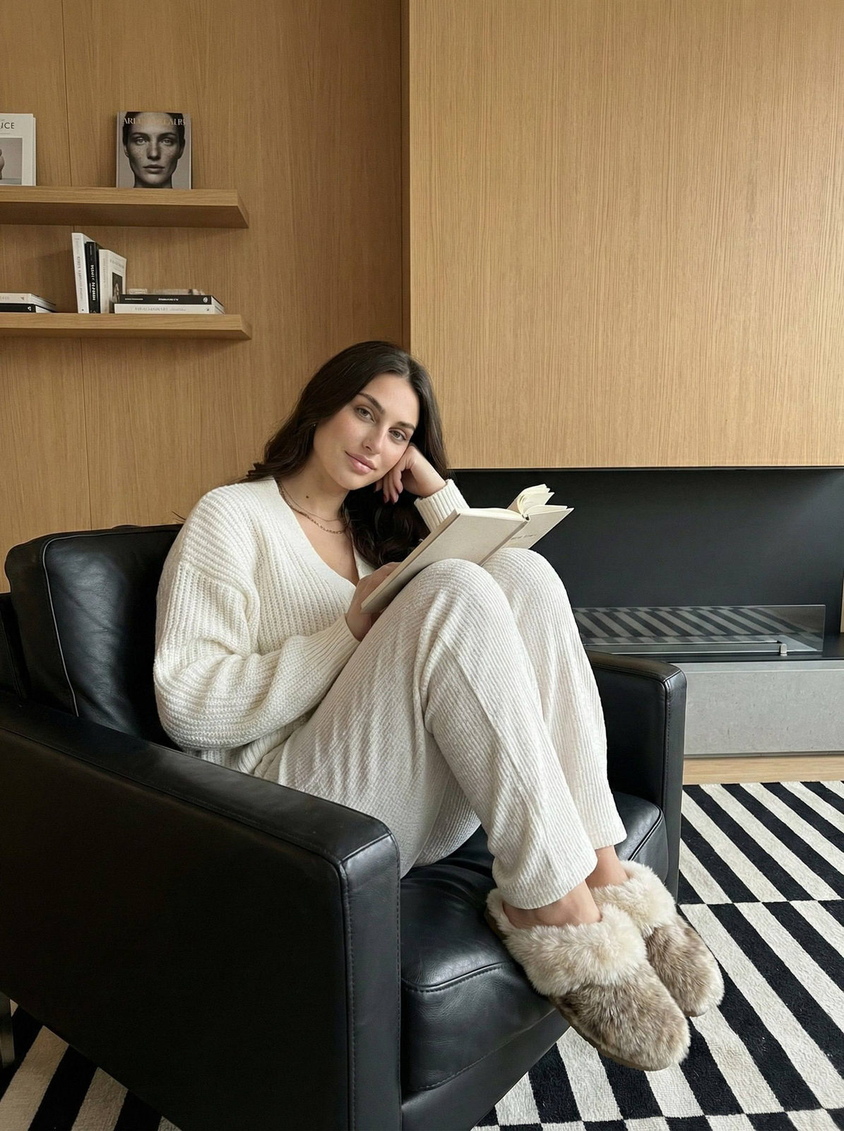 A young woman in a cozy setting sitting in a black leather chair, holding a book.