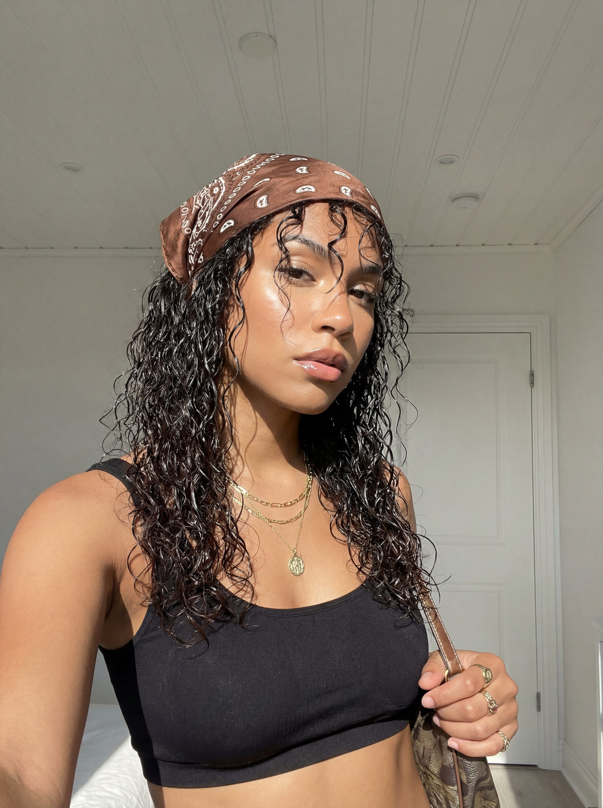 A woman in a paisley bandana and black top with glossy skin and layered necklaces, illuminated by soft sunlight.