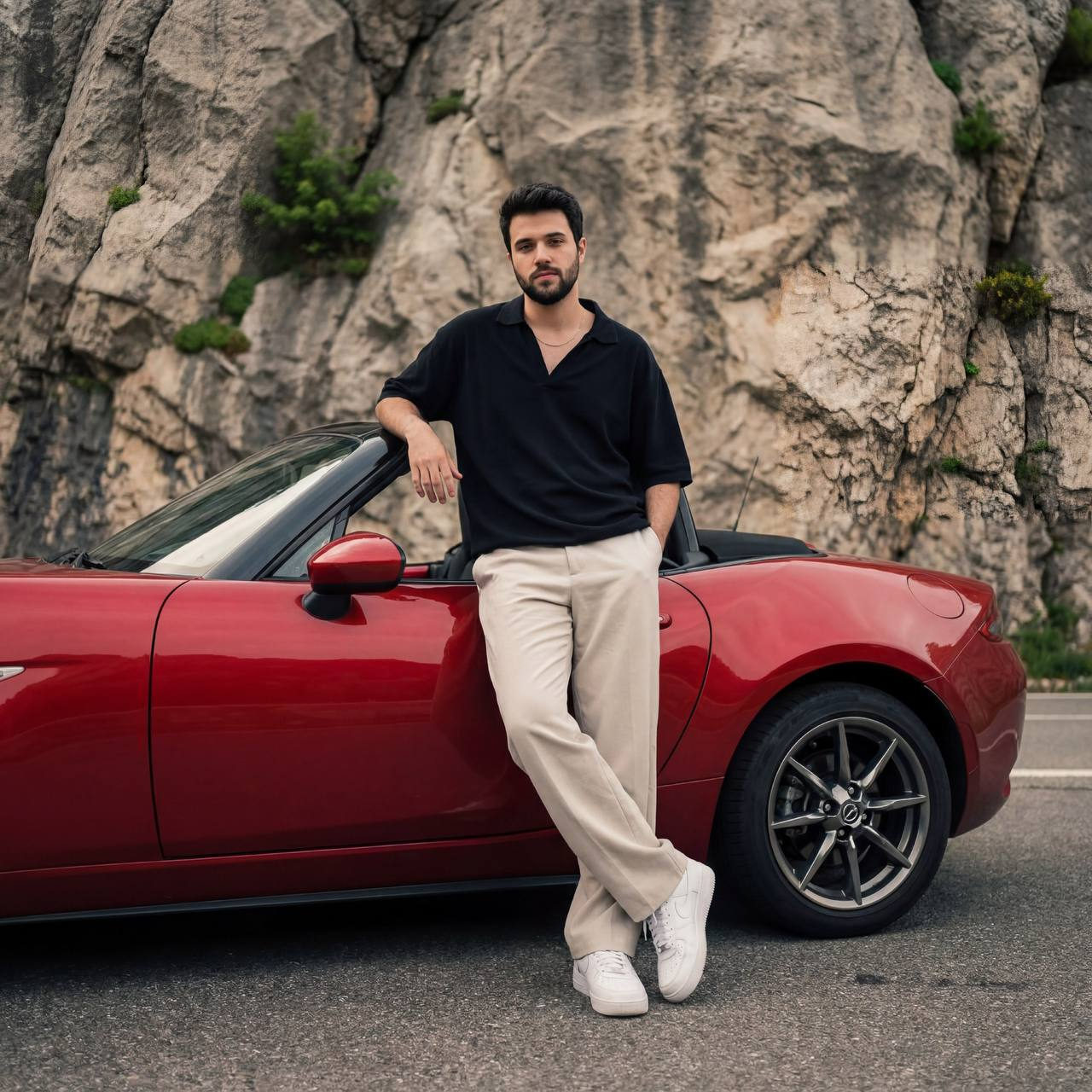 A young man in a loose black shirt casually leans against a red convertible, displaying a relaxed pose against a rocky cliff backdrop.