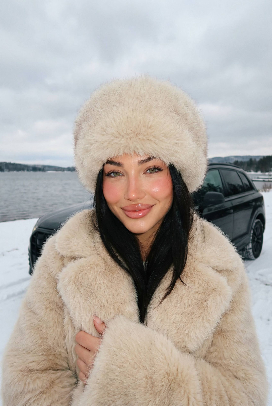A young woman in a cream fur coat smiling softly at a snowy lakeside.