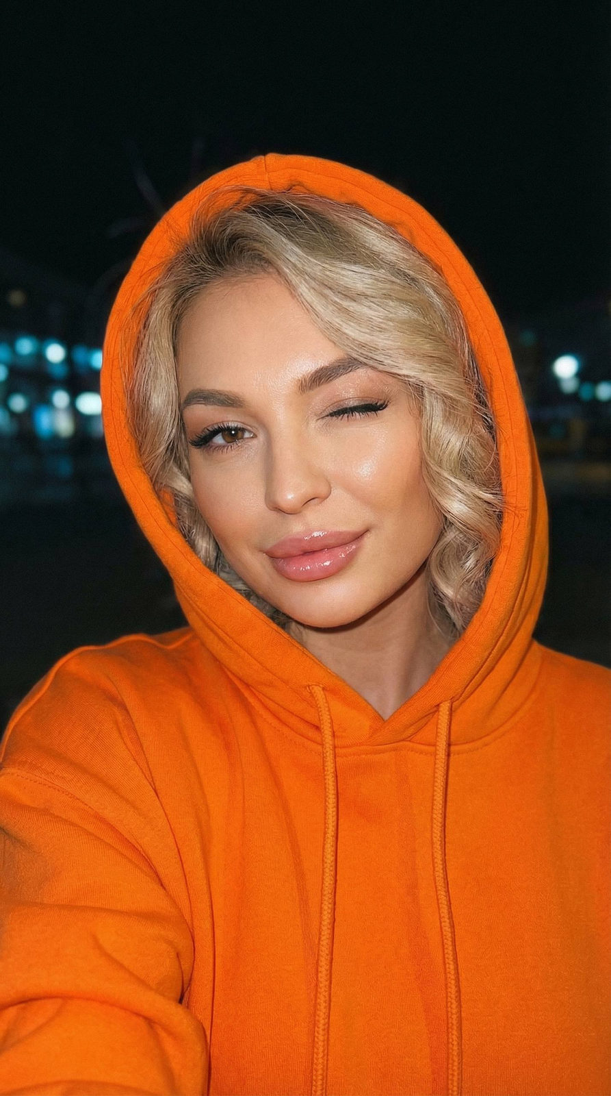 Close-up portrait of a woman in an orange hoodie winking with soft lighting and dark background.