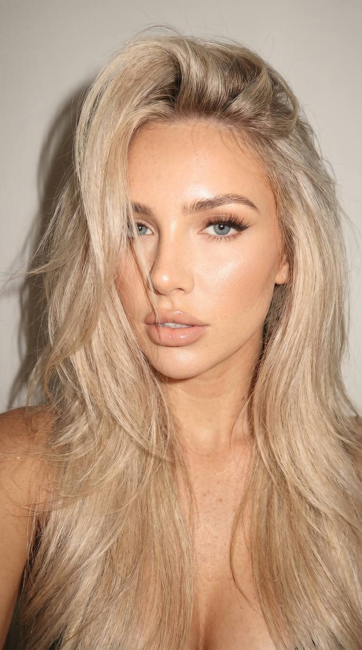 Photorealistic selfie portrait with voluminous blonde hair against a neutral wall, showcasing a candid expression and warm lighting.