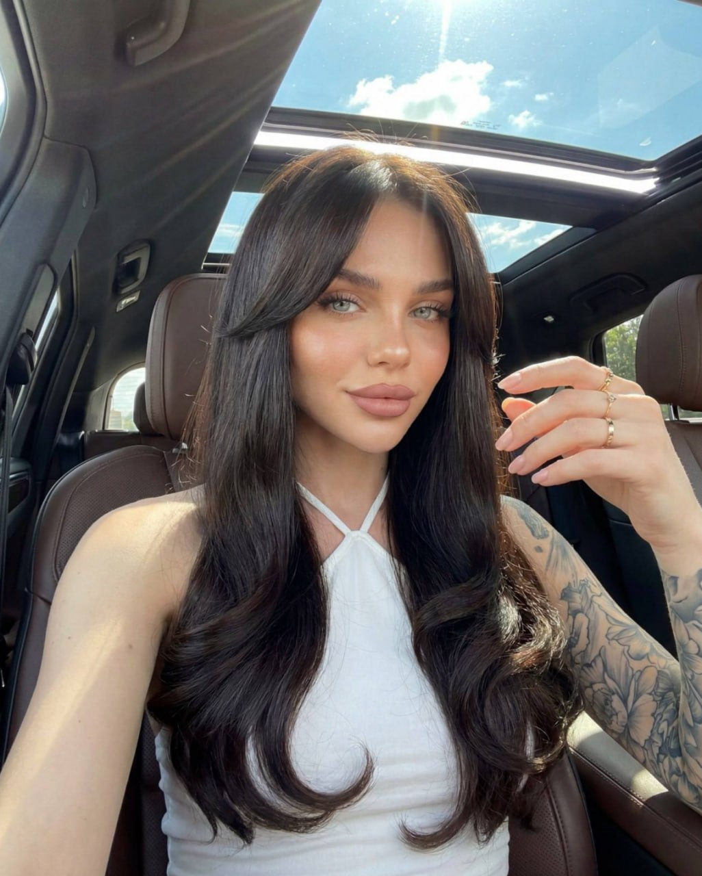 A woman in a luxury car, taking a selfie with an outstretched arm and confident smile.