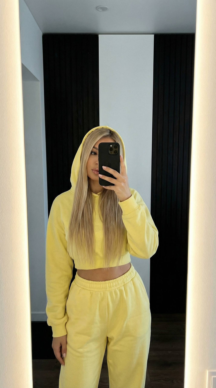 A woman taking a selfie in a mirror, wearing a lemon hoodie, with the reflection illuminated by side white light.