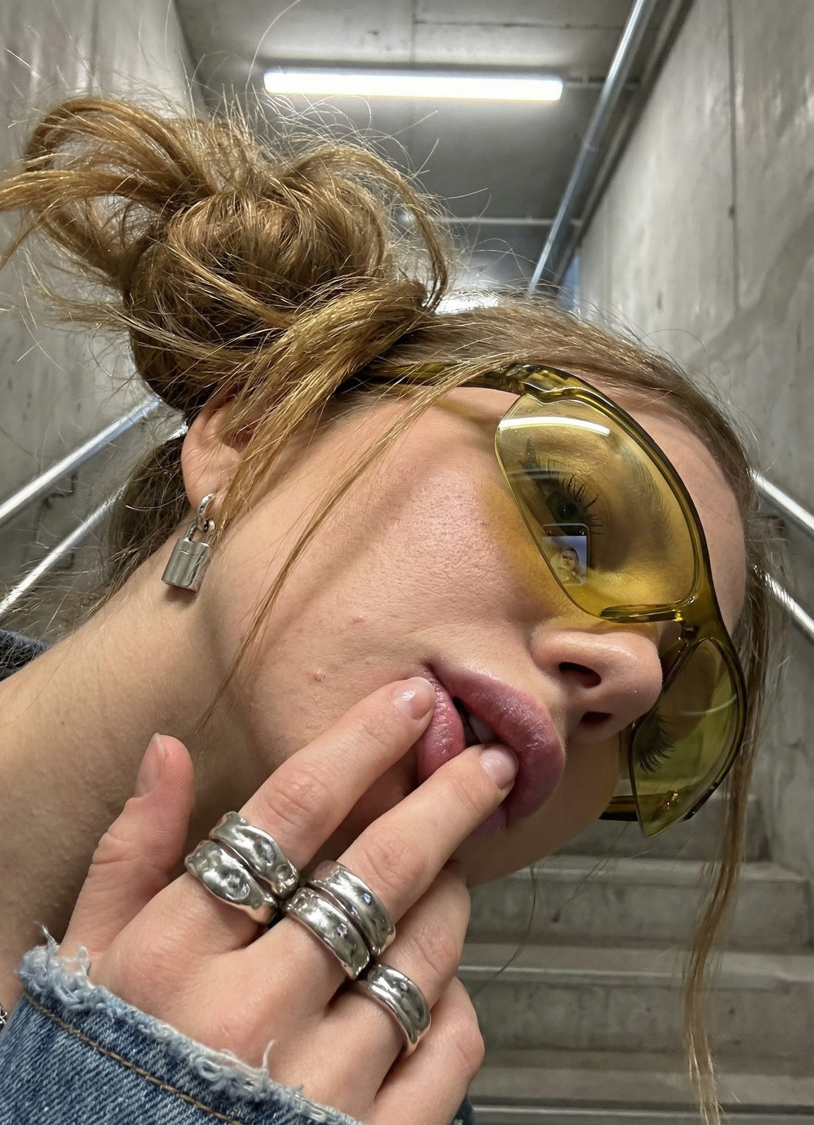 Close-up of a woman with messy bun and silver rings, showcasing frosted lips and golden strands in a stairwell light.