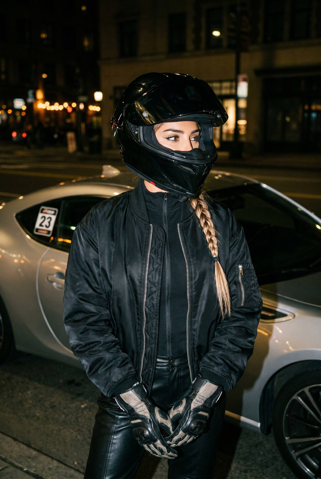 A woman in motorcycle gear stands before a silver sports car at night, hands holding racing gloves with a city street backdrop.