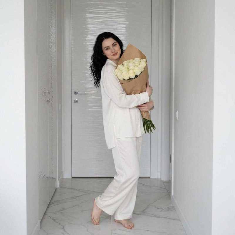 A woman in loose pajamas standing in a corridor, holding a large bouquet of roses, with a background of minimalist decor.