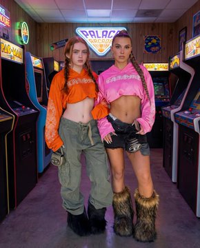 A woman in a bubblegum pink cropped hoodie and olive green cargo trousers stands in front of glowing arcade cabinets, wearing shaggy faux-fur boots.