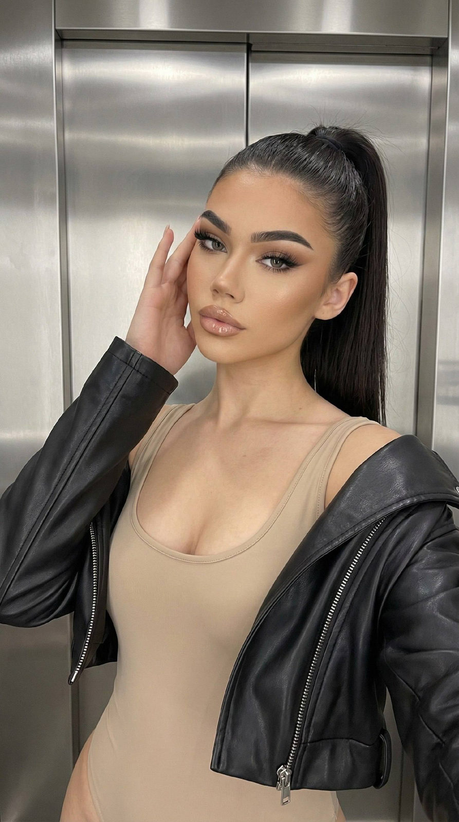 A stunning young woman poses confidently in an elevator, showcasing her high ponytail and glam makeup against a metallic wall.