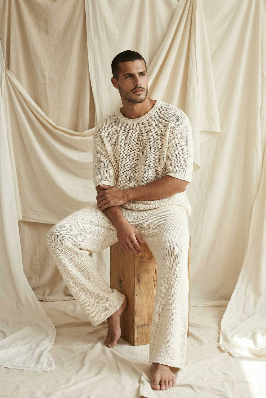 A male model in a minimalist studio wearing a loose-knit cream top and wide-leg trousers, seated on a wooden block against an ivory backdrop.