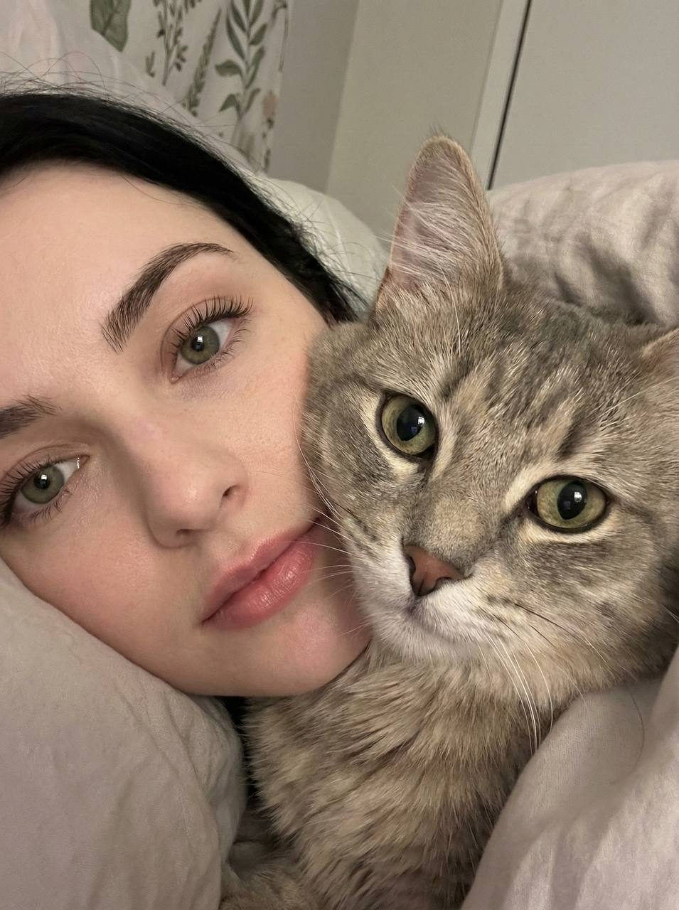 Close-up of a girl with long eyelashes and a calm gaze, with a fluffy gray tabby cat pressed against her face.