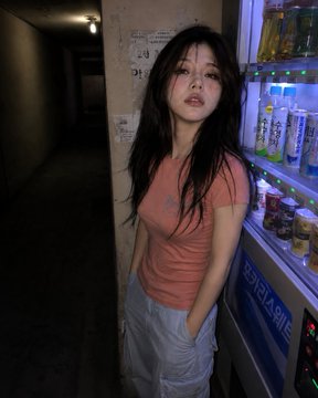 A woman in a peach shirt and blue cargo pants leans against a glowing vending machine in a dim corridor.