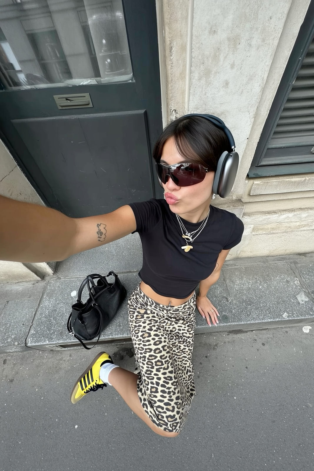 A candid wide-angle iPhone selfie of a young woman on a city ledge, wearing a black baby tee and leopard slip skirt, with vibrant yellow sneakers.