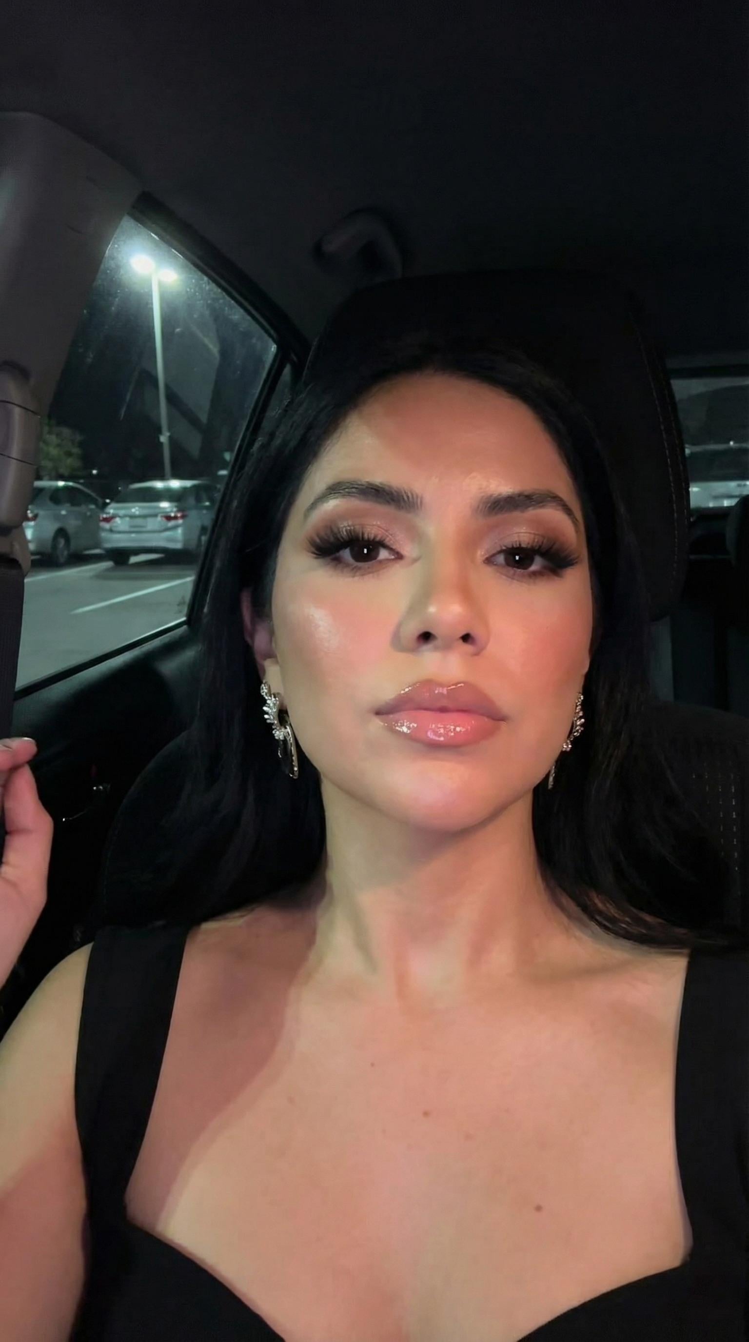 Close-up selfie of a young woman with glam makeup in a parked car at night, illuminated by a street lamp.