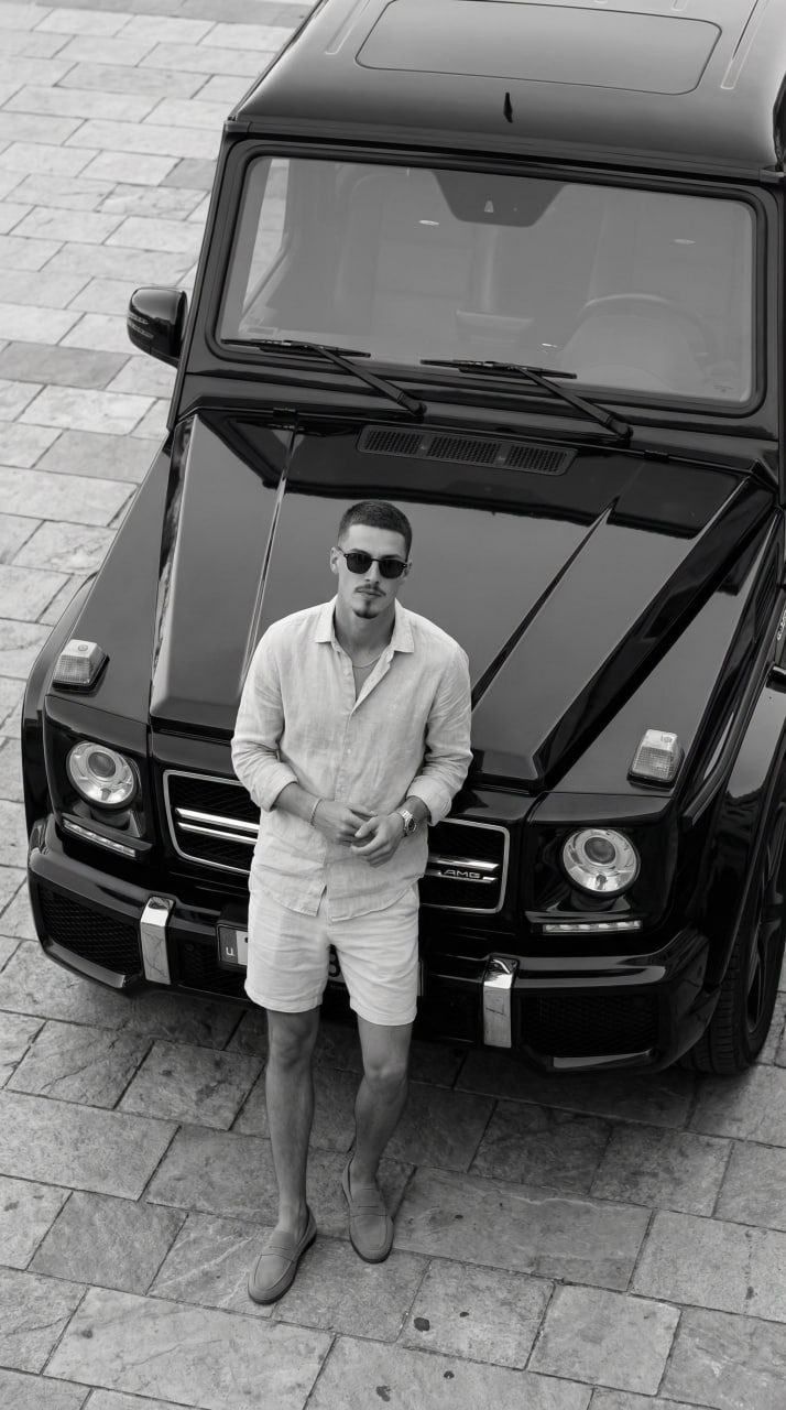 Monochrome portrait of a subject leaning beside a black Mercedes-Benz G 63 AMG on patterned stone pavement.