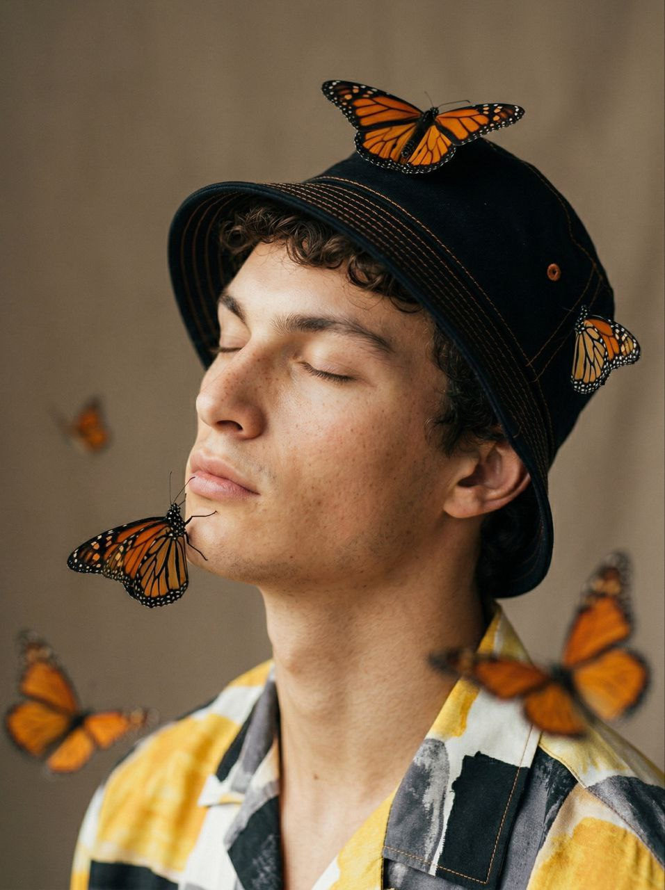 Close-up portrait of a young man wearing a bucket hat with orange stitching, surrounded by Monarch butterflies.