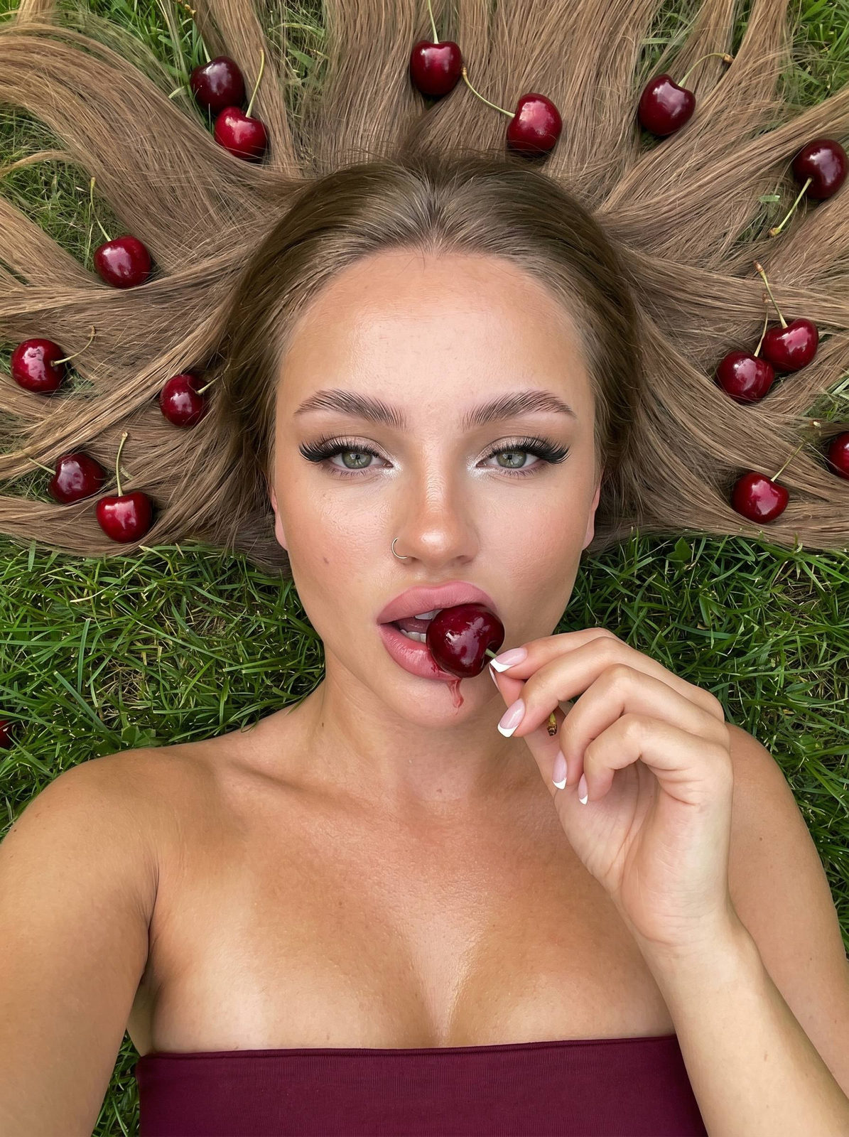 A woman in a burgundy top lying on fresh green grass, eating a large cherry with a passionate expression.